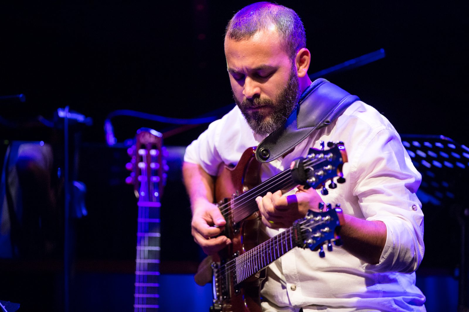 Cenk Erdoğan playing a double-neck guitar on stage under purple lighting.