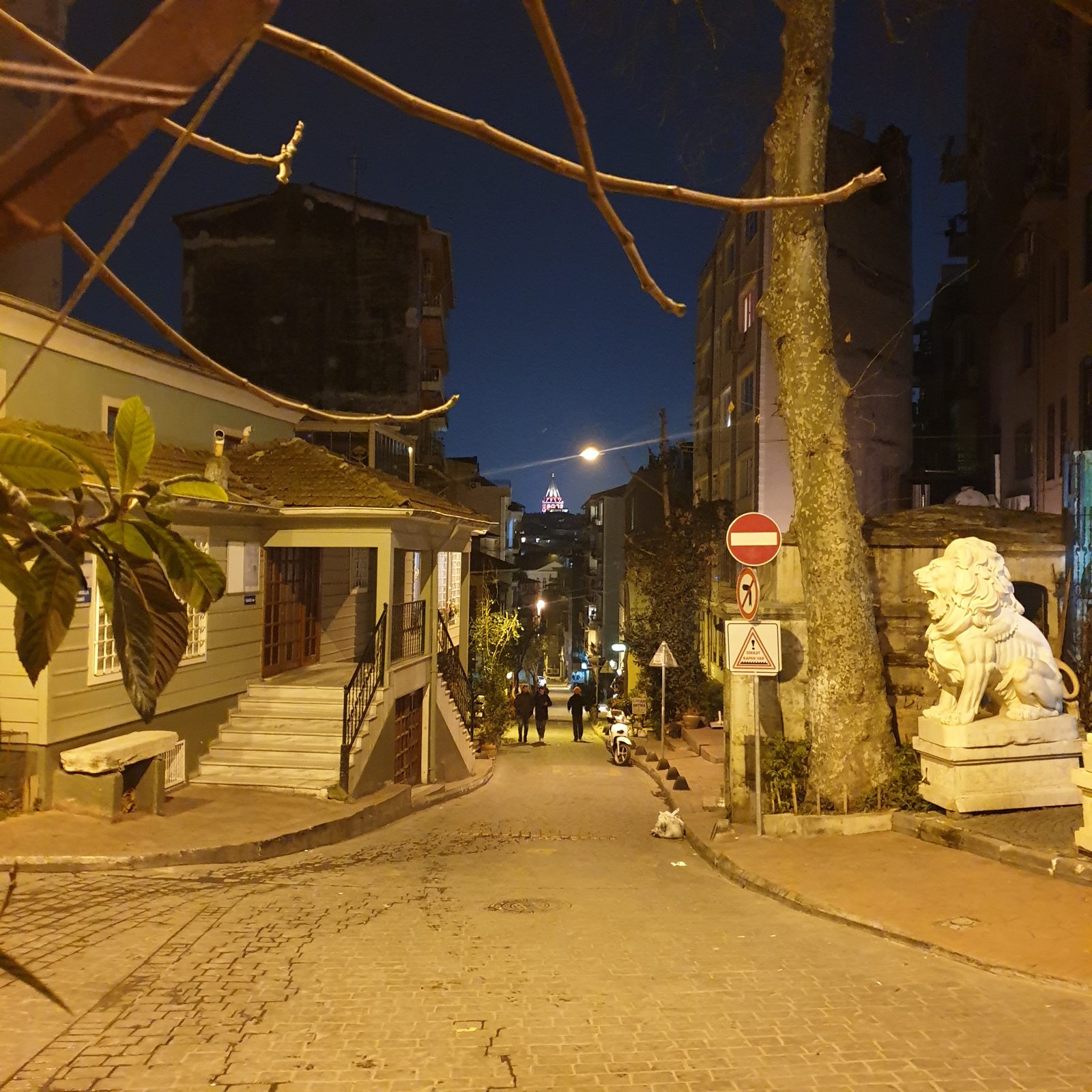 An evening view along Çukurcuma Caddesi, with shopfronts and Galata Tower visible in the distance.